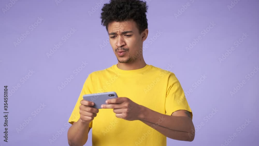 Young african emotional concentrated guy isolated over purple background play games by mobile phone Young african emotional concentrated guy isolated over purple background play games by mobile phone