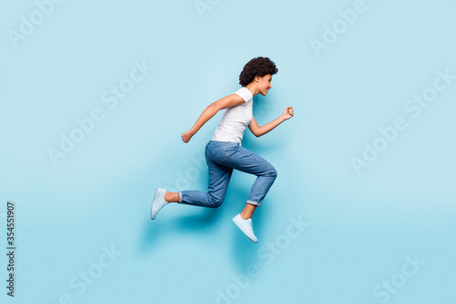 Full size photo of beautiful dark skin curly lady jumping high rushing marathon finish line coming first active sporty person wear casual white t-shirt jeans isolated blue color background