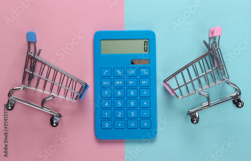 Two mini supermarket trolleys with calculator on blue pink pastel background. Shopping concept. Top view. Minimalism