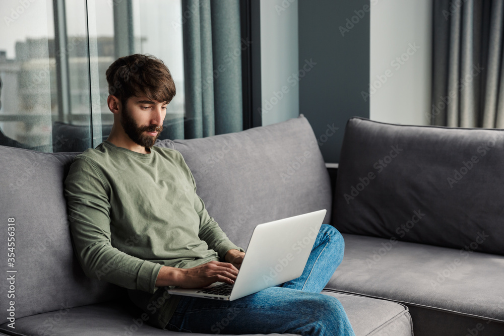 Image of focused young man using laptop while sitting on sofa Image of focused young man using laptop while sitting on sofa
