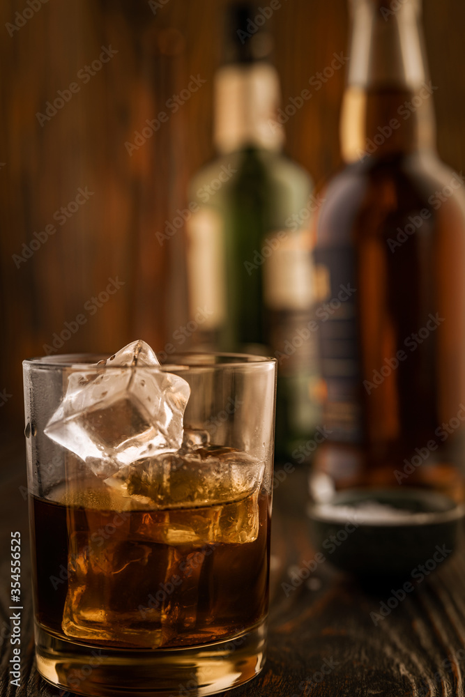 A glass of whiskey with ice, in the background are bottles on a wooden table of a bar counter, shallow depth of field, selective focus. The concept of alcoholic drinks in a roadside bar. A glass of whiskey with ice, in the background are bottles on a wooden table of a bar counter, shallow depth of field, selective focus. The concept of alcoholic drinks in a roadside bar.