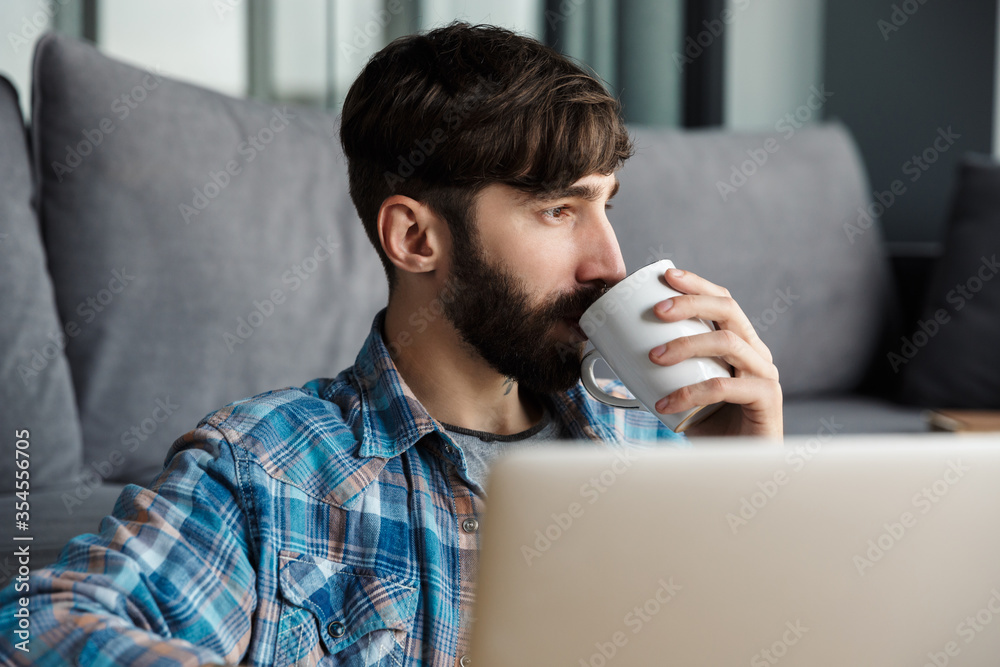 Image of man drinking coffee and using notebook while sitting on floor