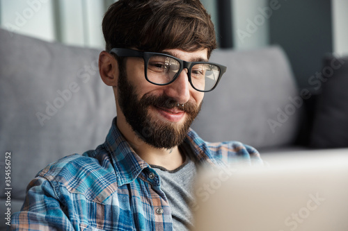 Image of happy bearded man in eyeglasses smiling and using notebook