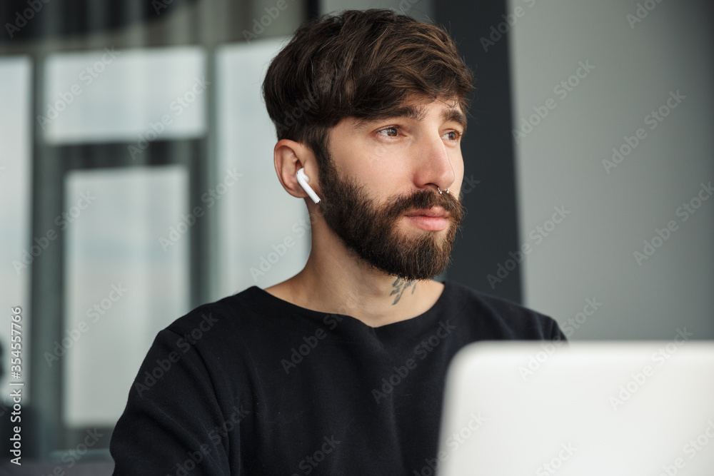 Image of serious man using wireless earphones and working with laptop Image of serious man using wireless earphones and working with laptop