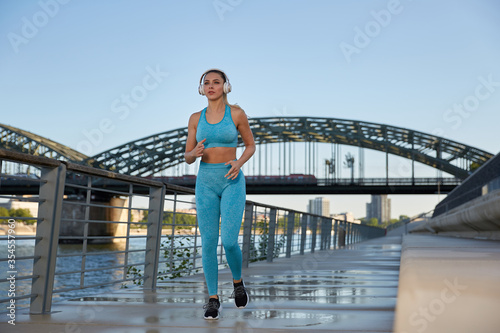 Fototapeta Naklejka Na Ścianę i Meble -  
Early in the morning, after sunrise, in the city by the river, a young attractive woman goes jogging on the promenade and listens to music through headphones