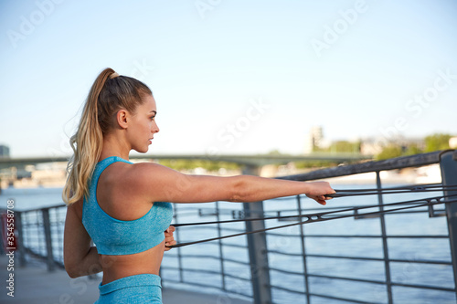 Fototapeta Naklejka Na Ścianę i Meble -  Early in the morning, after sunrise, in the city by the river, a young attractive woman makes fitness workouts on the promenade, outdoor and functional strength training with an elastic band, 