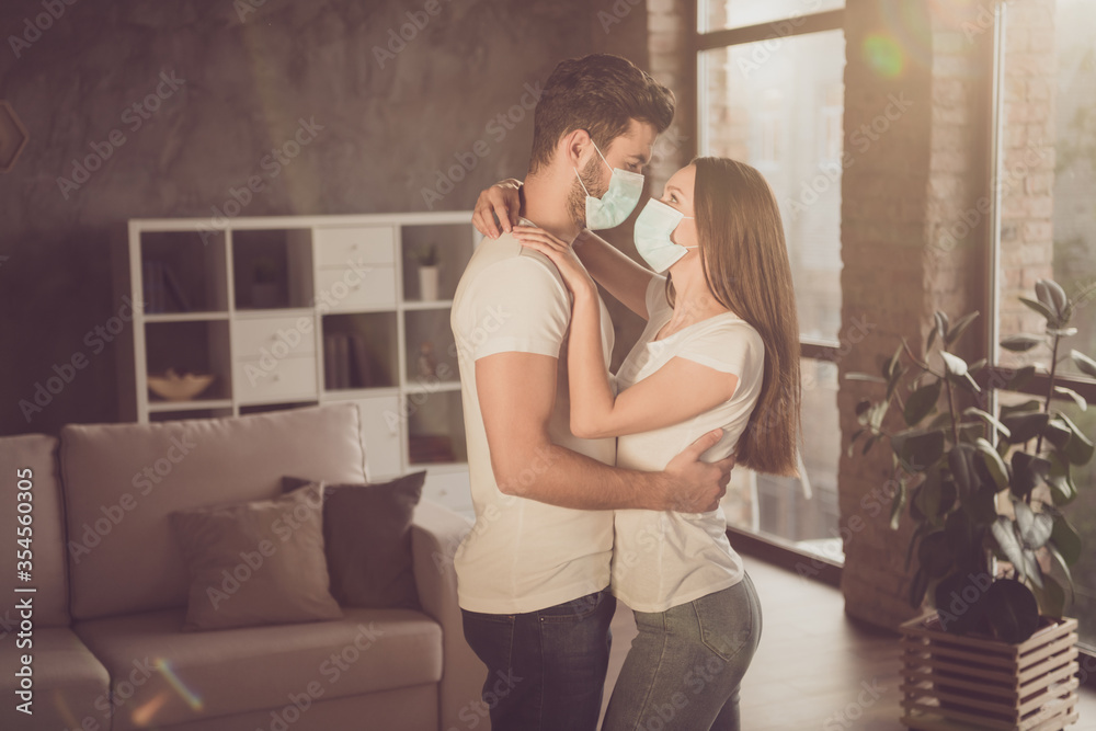Photo of young couple lady guy spend quarantine isolation together stay home hugging happy emotions stand opposite look eyes cozy wear facial medical mask living room evening sun indoors Photo of young couple lady guy spend quarantine isolation together stay home hugging happy emotions stand opposite look eyes cozy wear facial medical mask living room evening sun indoors