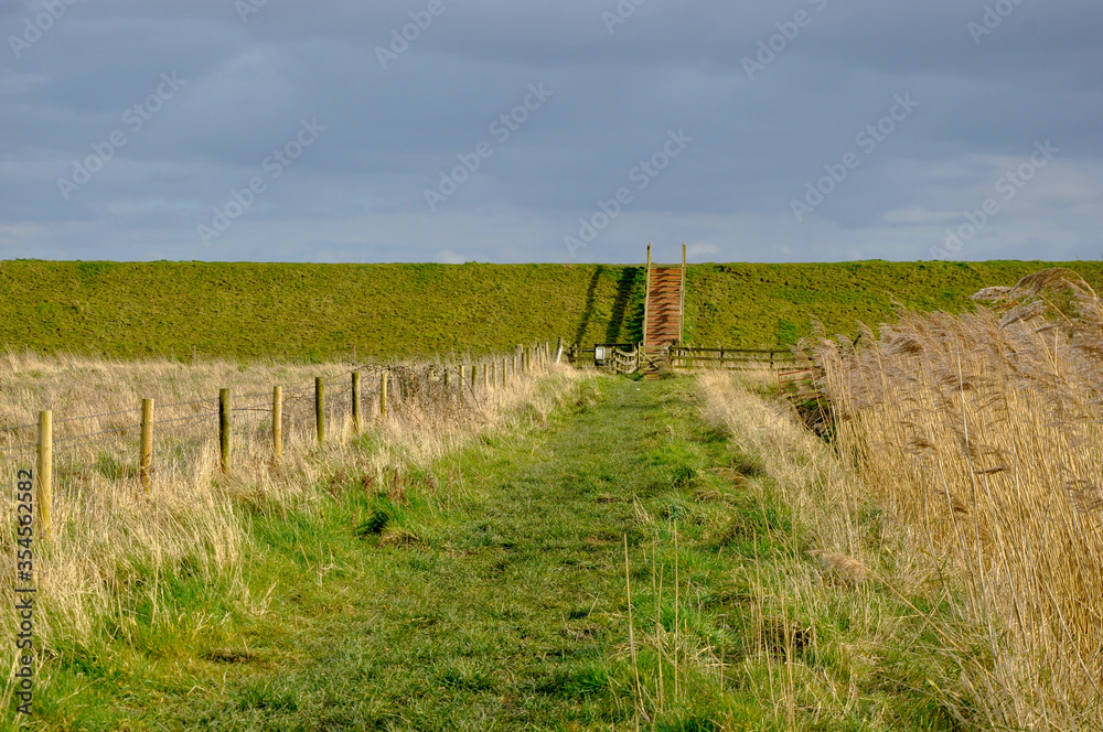 Wooden fence designates pathway across a nature reserve in Lincolnshire Fens towards steps over the sea wall into gathering clouds