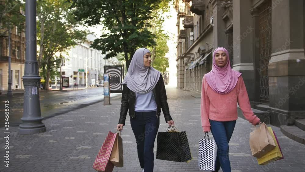 Two muslim dark-skinned females in casual clothes and colorful hijabs. They smiling and talking, walking down the street holding shopping bags