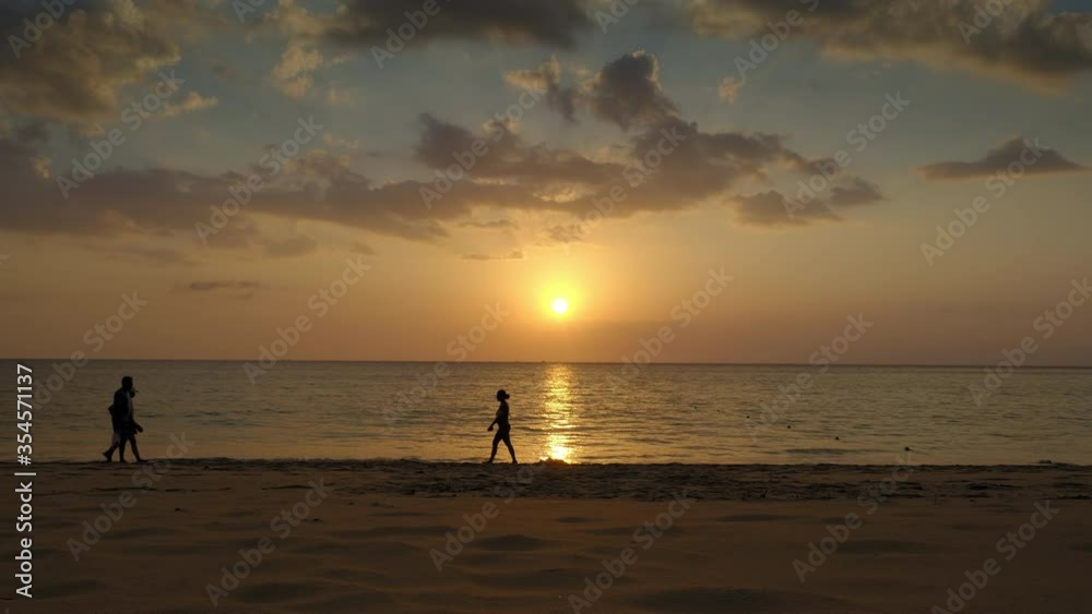 Silhouettes of people are walking in sunset beach with beautiful blue sky, yellow sun and waves in background Silhouettes of people are walking in sunset beach with beautiful blue sky, yellow sun and waves in background