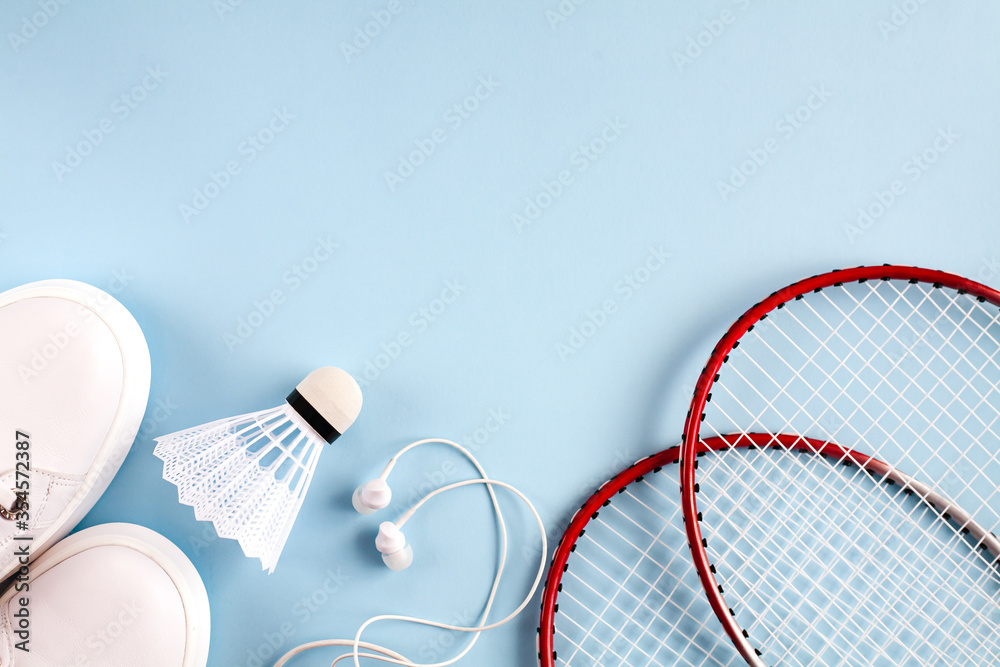 Sport flat lay with white shuttlecock, two rackets for playing badminton, white sneakers and white headphones on a blue background. Concept healthy lifestyle. Top view. Copy space.