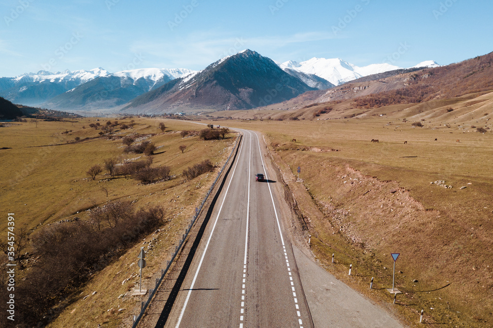 Aerial view of A-155 Sukhumi Military Road with moving car, brown autumn grass, grazing horses, high snow-capped mountains and clear blue sky in the background, the Caucasus