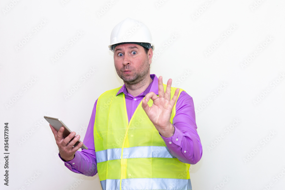 Construction builder wearing helmet yellow protective vest using wireless tablet and showing like or thumbs-up shocked poke up shows to you and victory gesture isolated on white background. Construction builder wearing helmet yellow protective vest using wireless tablet and showing like or thumbs-up shocked poke up shows to you and victory gesture isolated on white background.