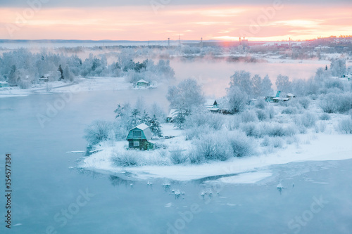 Cold orange dawn with gentle fog over wooden dacha houses on Angara River island, snowy trees and frozen water, Irkutsk, Siberia