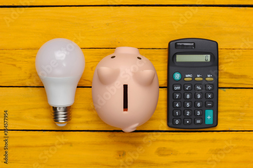 Saving electricity. Led light bulb and piggy bank, calculator on yellow wooden background. Top view