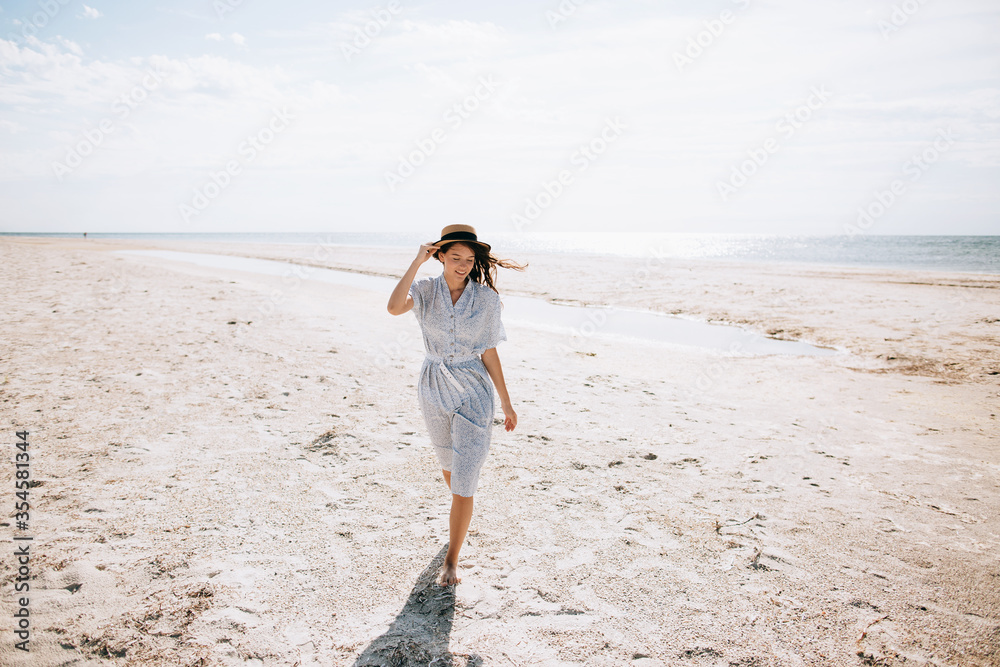 Lonely happy young woman in a hat and dress walks on a deserted sea beach on a sunny day. Summer vacation
