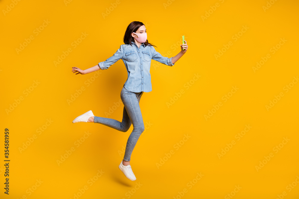 Full length photo of beautiful girl jump use smartphone make selfie video call connection technology wear medical mask denim jeans shirt isolated over yellow color background
