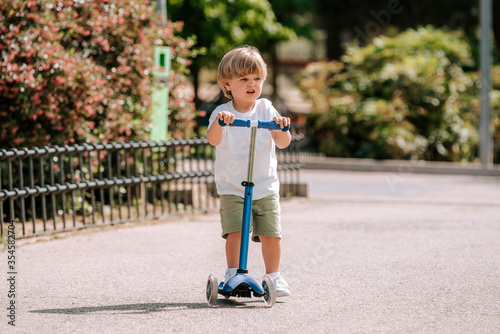 Boy learn to ride scooter in a park on sunny summer day. Preschooler boy riding a roller. Kids play outdoors with scooters.