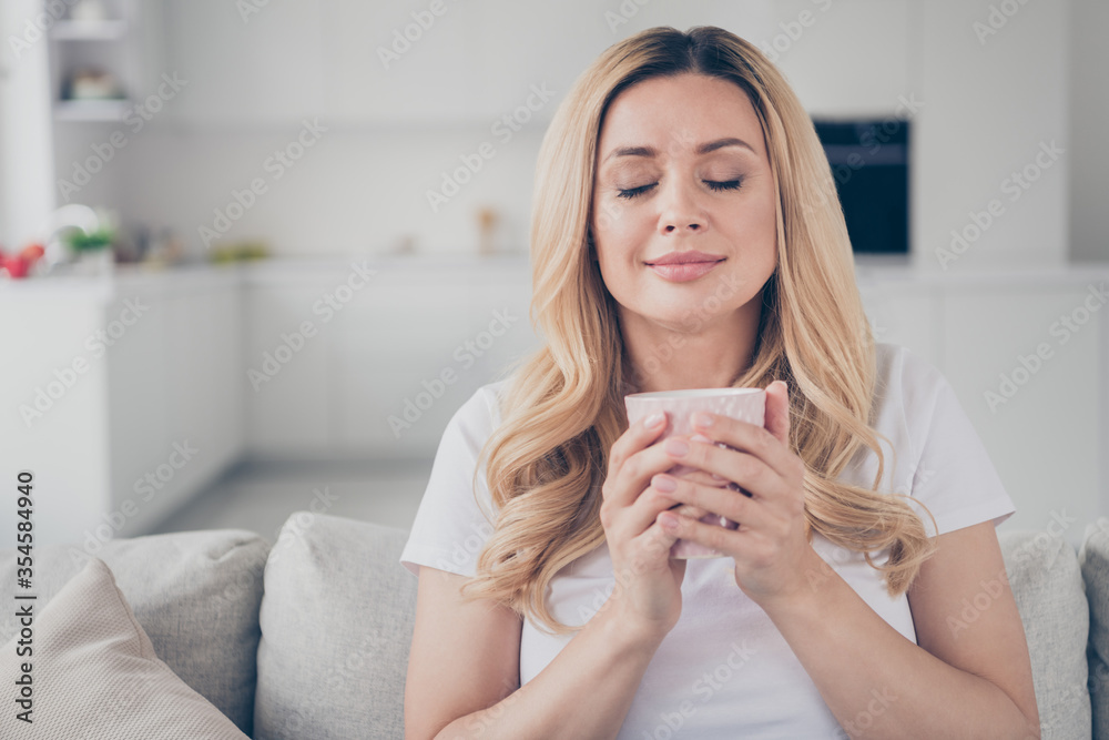 Closeup photo of domestic cheerful blond lady eyes closed relaxing sit comfy couch holding hot beverage mug dreamer enjoy quarantine stay home good mood living room indoors