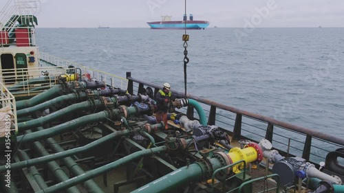 concentrated man in white protective helmet stands aboard of Natig Aliev tanker connecting product pipelines against container ship in sea photo