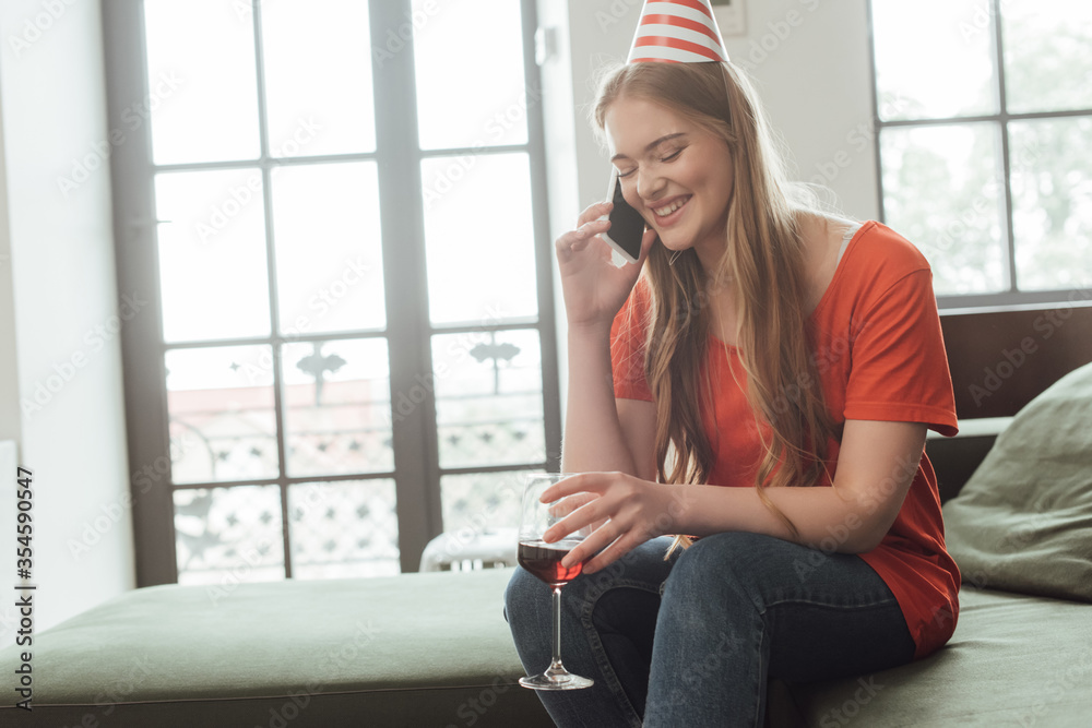 cheerful girl in party cap holding glass of red wine and talking on smartphone at home cheerful girl in party cap holding glass of red wine and talking on smartphone at home