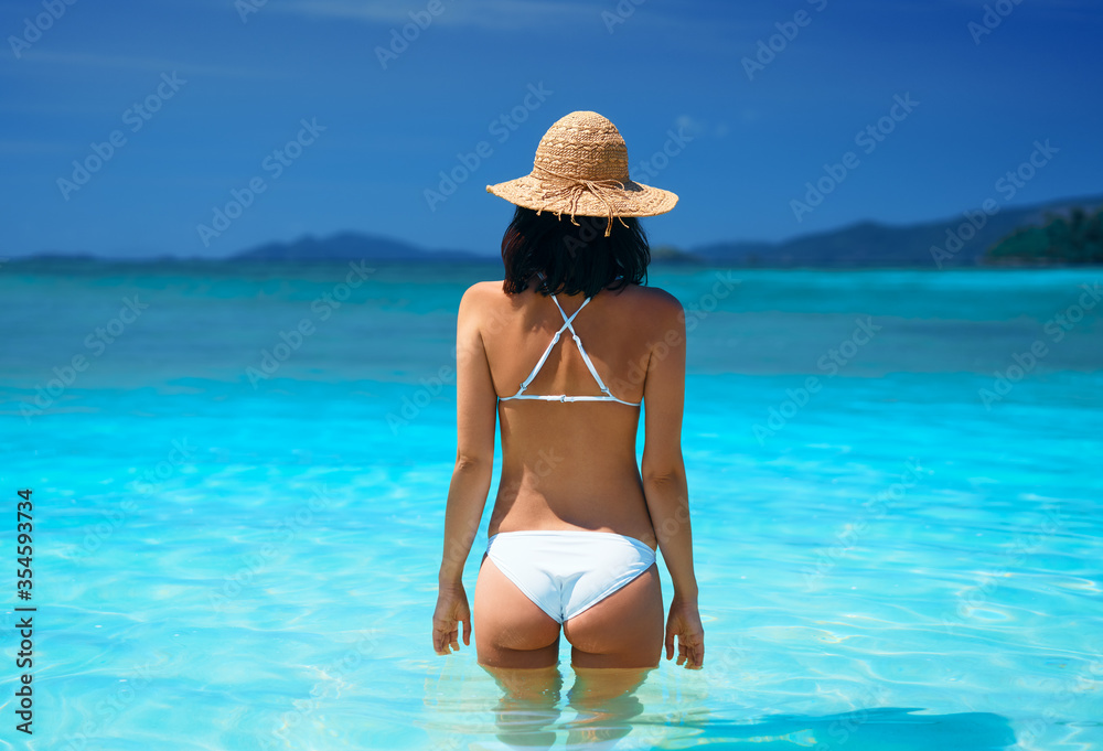 Back view of young sexy woman in white bikini and straw hat relax in turquoise sea water on tropical beach in paradise island