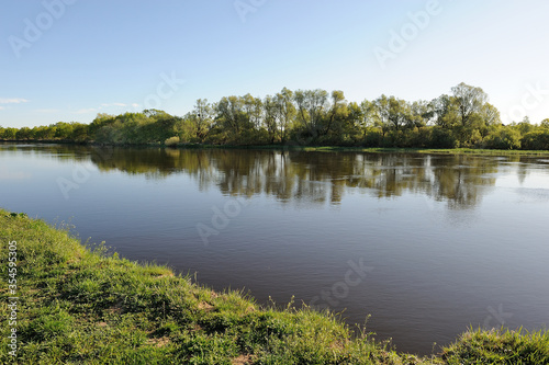 Summer. Morning landscape with a flat river, green grass and a cloudless blue sky