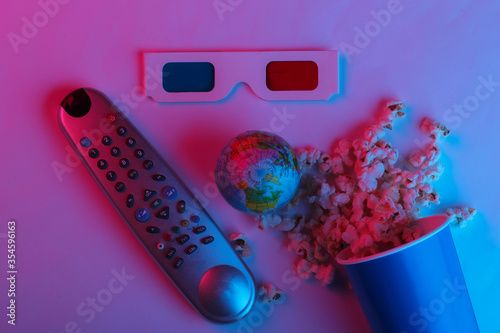 ovie time. Cardboard bucket of popcorn, tv remote and Stereoscopic anaglyph disposable paper 3d glasses, globe in pink blue gradient neon light. Top view photo