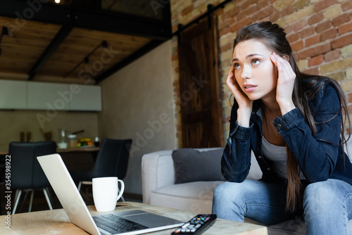 selective focus of concentrated girl touching head while watching movie near laptop and cup