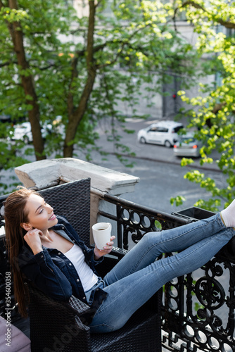 smiling girl in denim jeans and jacket sitting on chair and holding cup of coffee on balcony