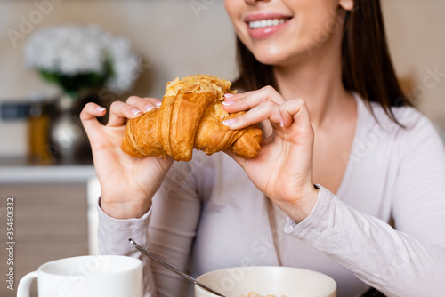 cropped view of happy girl holding tasty croissant near bowl and cup