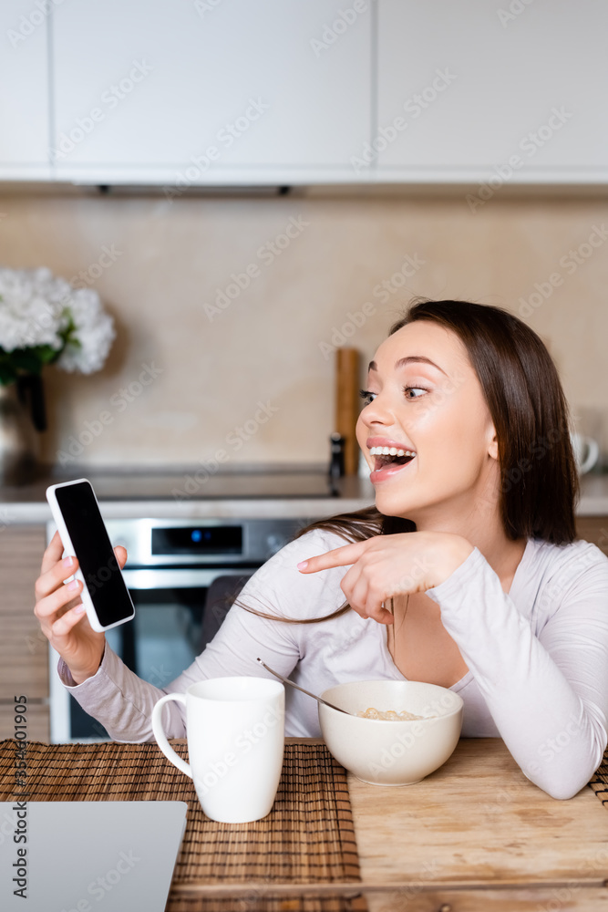 excited girl pointing with finger at smartphone with blank screen near cup and bowl excited girl pointing with finger at smartphone with blank screen near cup and bowl