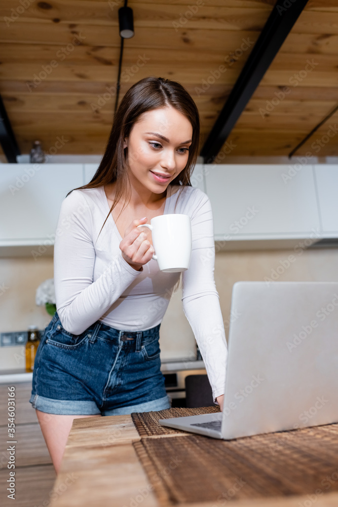 selective focus of happy freelancer using laptop and holding cup with tea