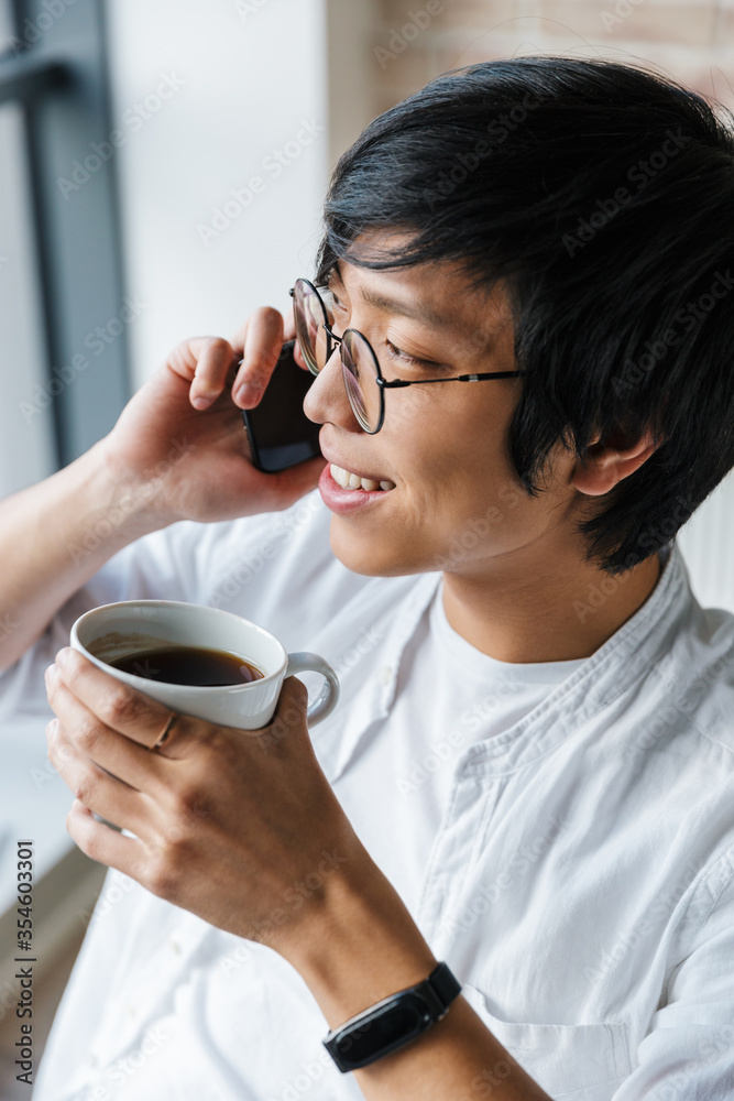 Image of young asian man using cellphone and drinking coffee in office Image of young asian man using cellphone and drinking coffee in office