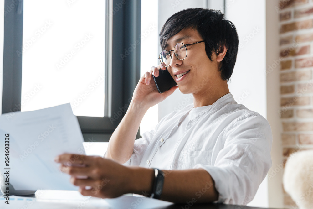 Image of asian man using cellphone and holding paper documents in office Image of asian man using cellphone and holding paper documents in office