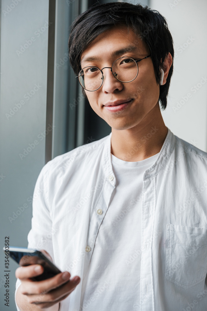 Image of young asian man wearing earphones using cellphone in office Image of young asian man wearing earphones using cellphone in office