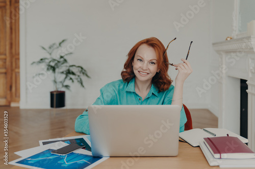 Indoor shot of happy redhead young European woman works on laptop computer, communicates online, sits in coworking space with paper documents, holds spectacles, wears blue shirt, browses web.