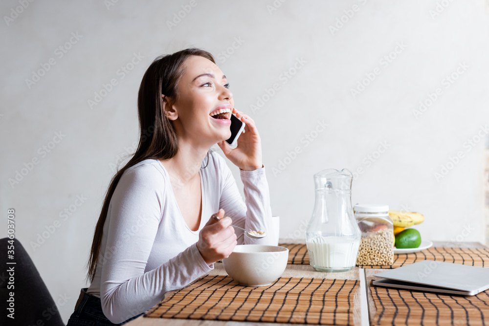 happy girl talking on smartphone and laughing while holding spoon with tasty corn flakes happy girl talking on smartphone and laughing while holding spoon with tasty corn flakes