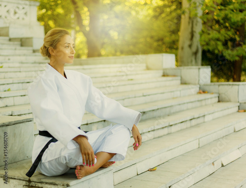 Portrait of young woman in white kimono with black belt. Sport woman sitting on stairs outdoors. Martial arts
