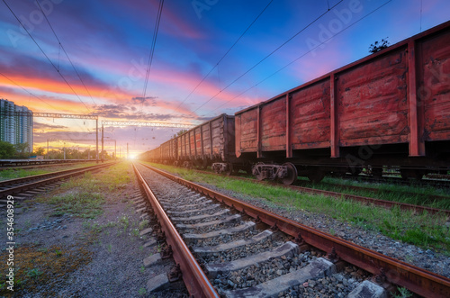 Railway station with freight trains at colorful sunset. Railroad in summer. Heavy industry. Industrial landscape with train, green grass, railway platform, blue sky with pink clouds. Transportation