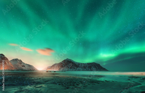 Aurora borealis above the snowy mountain and sandy beach in winter. Northern lights in Lofoten islands, Norway. Starry sky with polar lights. Night landscape with aurora, sea coast, city lights