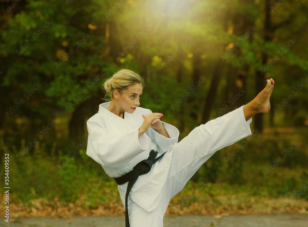Attractive female master fighter in white kimono with black belt kicks outdoors. .