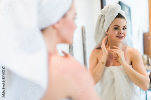 Beautiful smiling young woman in robe and towel touching face while looking at mirror in bathroom