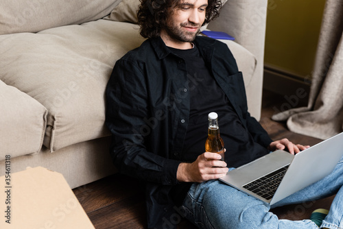 Selective focus of man holding beer bottle and using laptop near pizza box on floor
