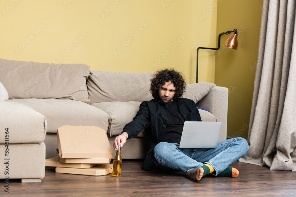 Freelancer holding bottle of beer near laptop and pizza boxes on floor in living room Freelancer holding bottle of beer near laptop and pizza boxes on floor in living room