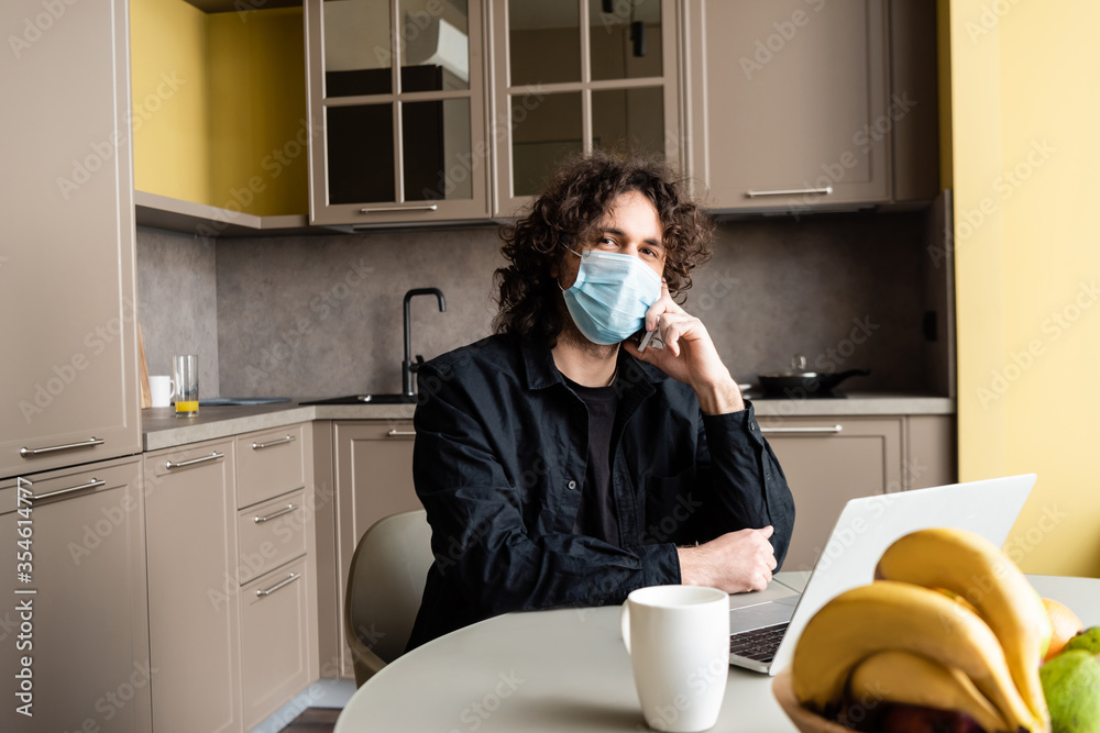 Selective focus of freelancer in medical mask talking on smartphone near laptop, fruits and cup on kitchen table Selective focus of freelancer in medical mask talking on smartphone near laptop, fruits and cup on kitchen table