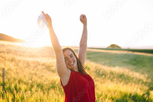 Happy smiling young woman, is standing with raised arms up, holding protective face mask in her hand. Woman enjoying breathing in nature outdoors. Celebrating end of Coronavirus pandemic .