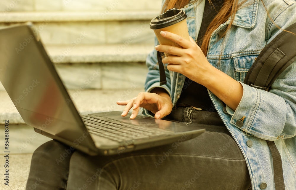 Fototapeta premium Happy modern woman student in denim jacket and backpack sitting on the stairs and uses laptop outdoor. Drinks coffee. Distance learning. Modern youth concept.