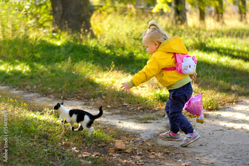 Сhild girl in a yellow jacket is trying to catch up with a black and white cat to pet.