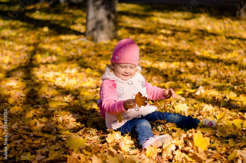 Little girl playing in autumn park. A girl sits in maple leaves and holds a sheet of maple in his hand.